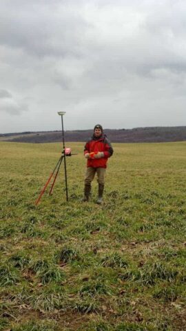 Surveyor standing in open field with surveying equipment under cloudy sky.