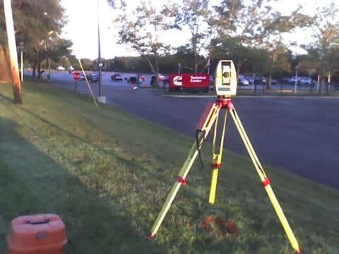 Land surveyor's tripod and equipment on grassy roadside near parking lot.