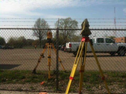 Two survey tripods with total stations behind a chain-link fence.