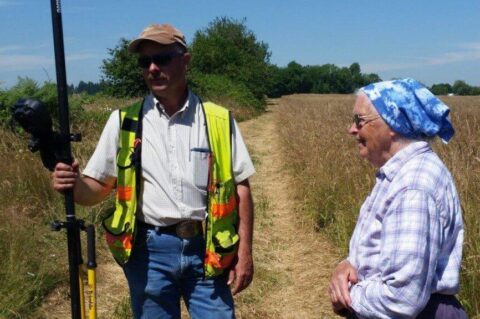 Surveyor with equipment and an elderly woman in a rural field.