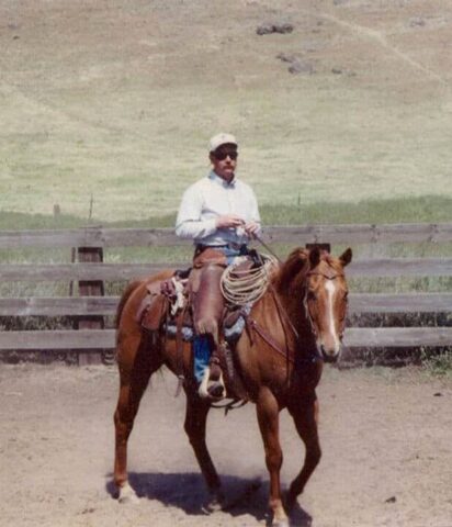 Man riding a brown horse in a rural open field with fencing and hills.