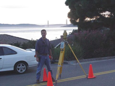 Surveyor with total station equipment near water with bridge in background.