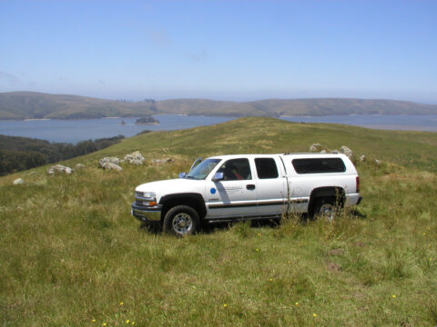 White survey vehicle on grassy hill with lake and hills in background.